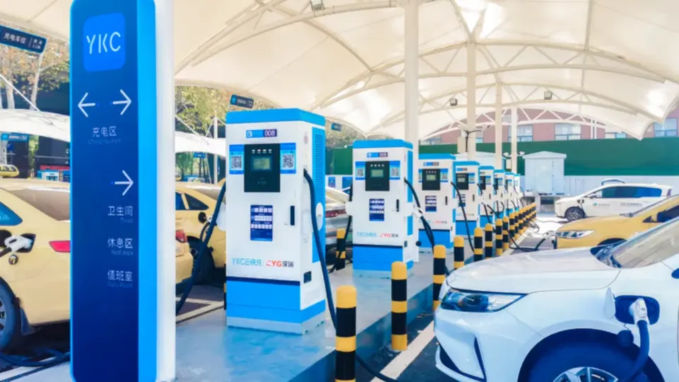 Electric car charging station row under a white canopy, blue-and-white charging kiosks with cars plugged in along a curbside lane.