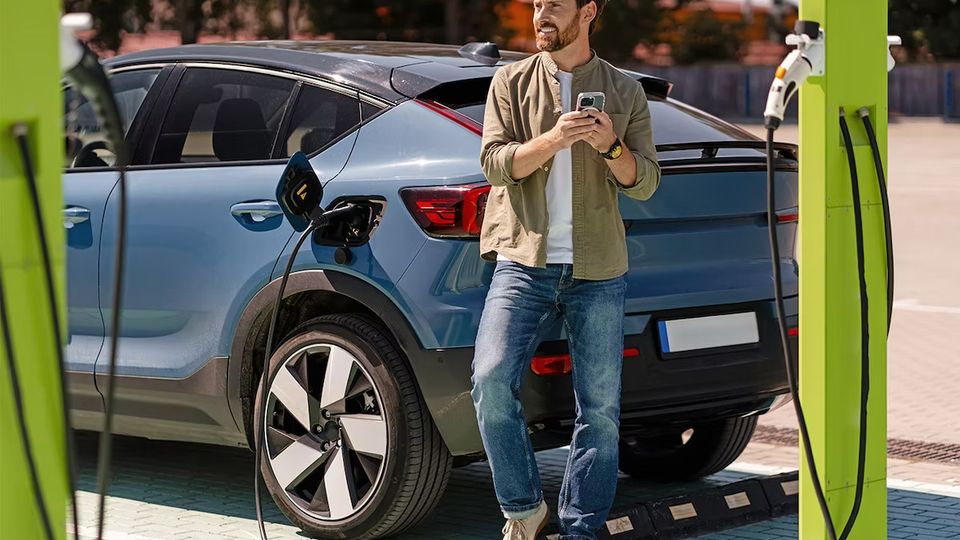 Man stands beside a blue electric SUV at a charging station, holding a phone while plugging in the charger.