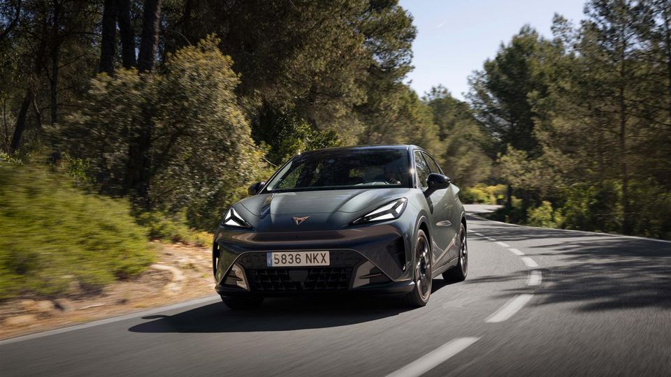 Front three-quarter view of a gray compact crossover driving on a winding forest road, sunlight filtering through trees, license plate 5836 NKX visible on the front bumper