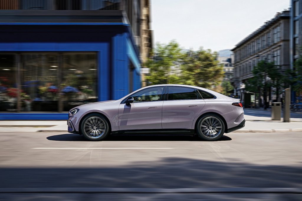 Silver luxury sedan driving along a city street past a blue storefront on the left and buildings on the right.
