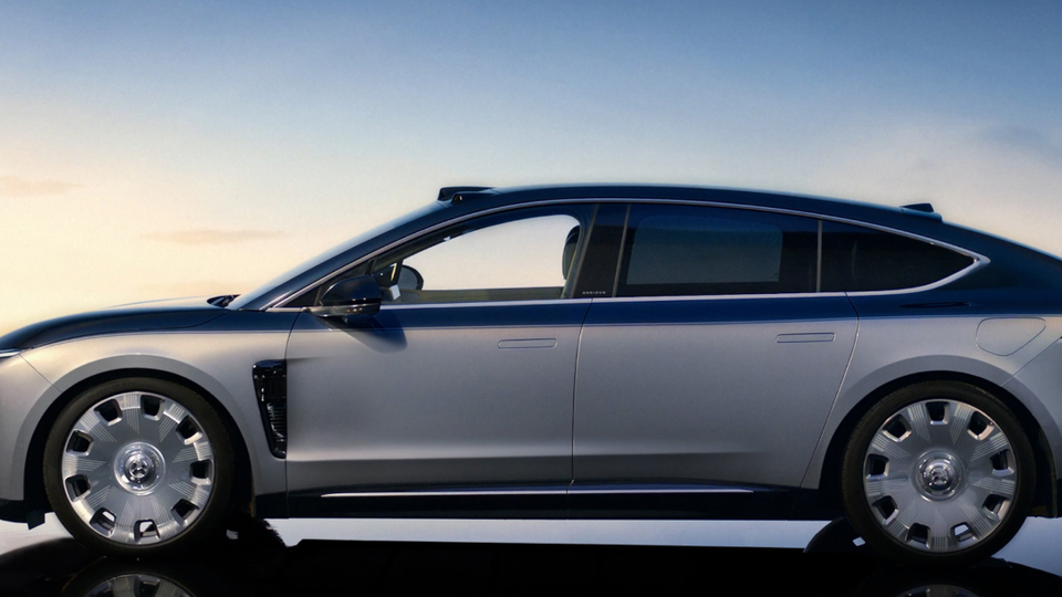 Side profile of a silver-blue futuristic sedan with large alloy wheels on a mirrored surface at sunset ground, showing a low, aerodynamic silhouette and flush door handles.