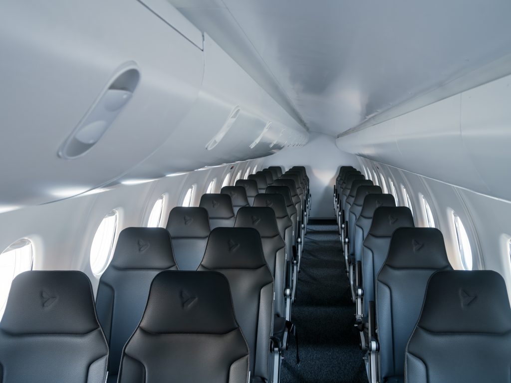 Interior of a passenger airplane cabin with multiple rows of dark gray seats and overhead storage, viewed from the aisle