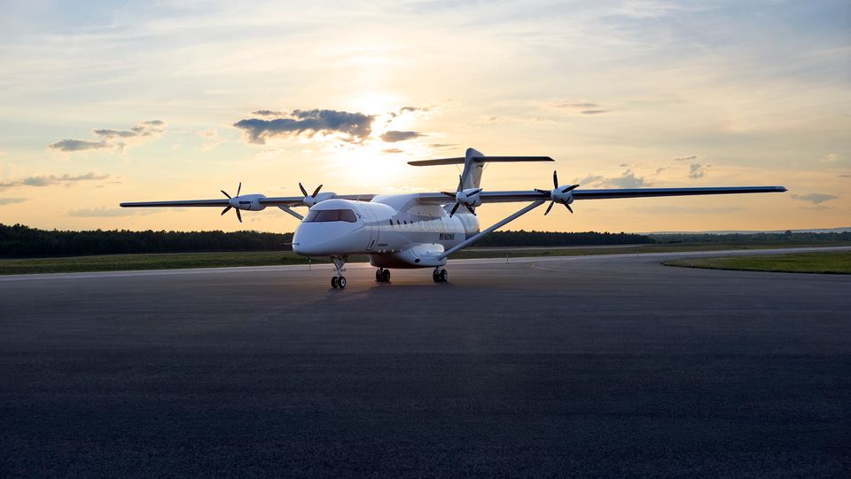 White twin-propeller airplane parked on a runway at sunset with a low sun near the horizon and trees in the distance.