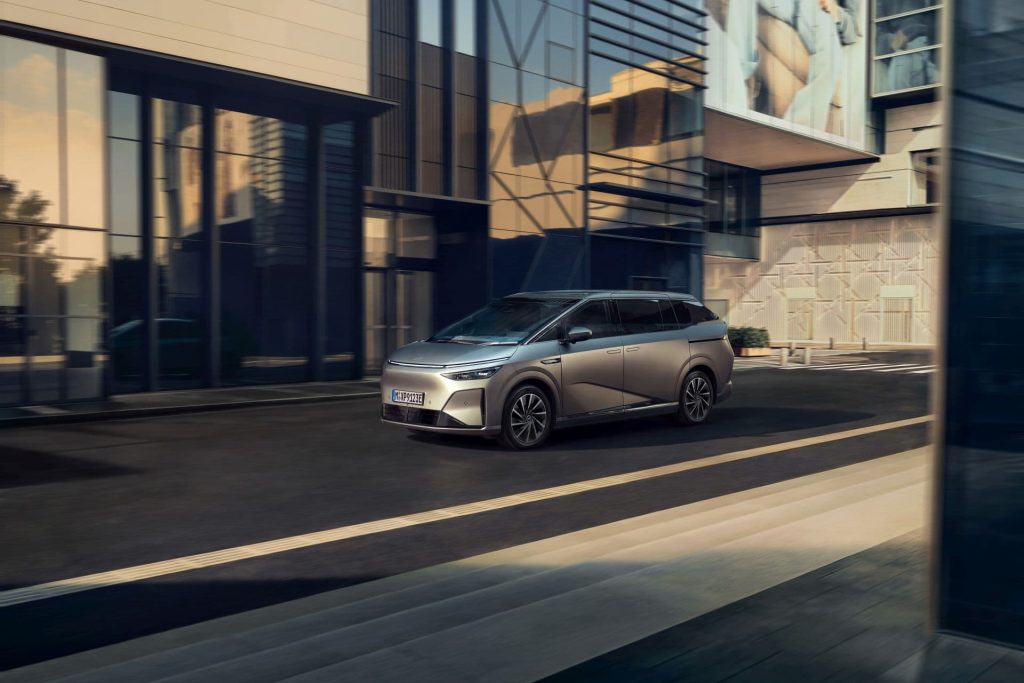 Silver minivan driving along a modern city street with glass-fronted buildings nearby.